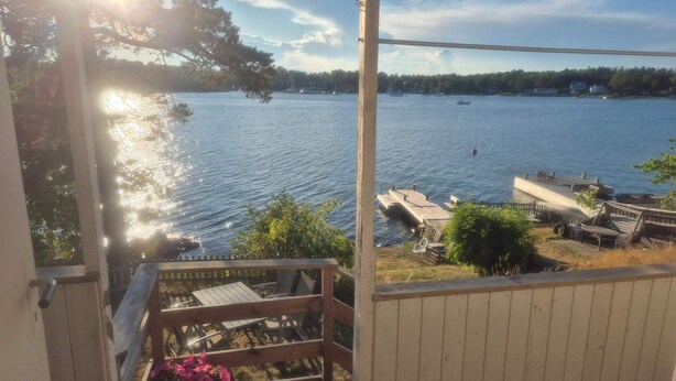 Une vue depuis un balcon donnant sur un lac, avec des bateaux, un quai et une terrasse en bois équipée de chaises et d’une table.