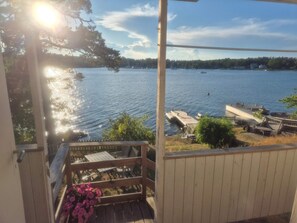Blick vom Balkon auf einen See mit Booten, einem Steg und einer Holzterrasse mit Stühlen und einem Tisch.