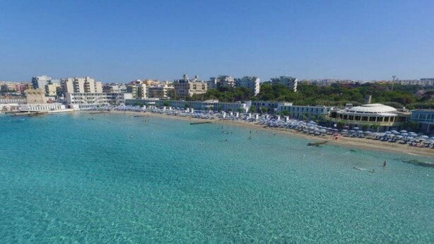 Une zone côtière dotée d’une plage, d’une eau turquoise et limpide, et d’une rangée de bâtiments situés en bord de mer, chacun agrémenté de parasols.