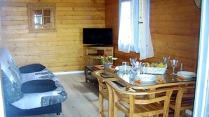 A wooden cabin interior with a sofa, a dining table set for four, a television, and a window with curtains. - CHALET JAUNE, a 2300m2 park, wifi, parking, swimming pool, kitchen, happiness (Saint-Pierre)