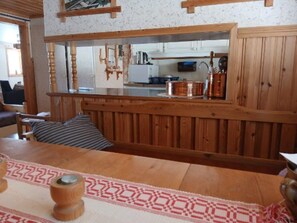 A wooden dining table with a patterned runner, a bench, and a kitchen area visible through a window.
