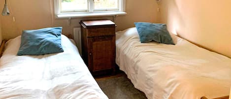 A bedroom with two single beds, a small wooden cabinet, a window with a view of greenery, and a shelf with books.