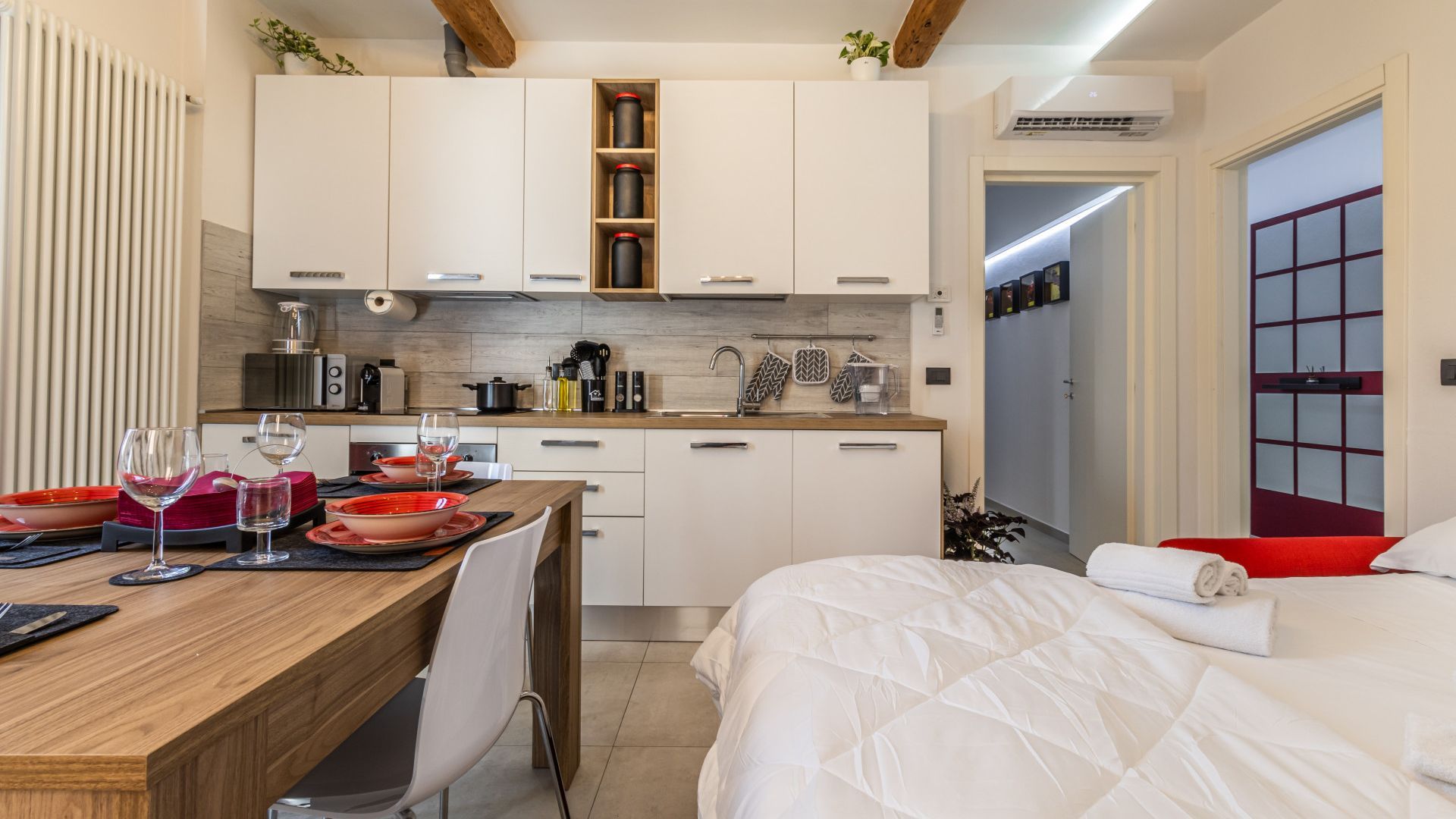 Une chambre d’hôtel compacte dotée d’une kitchenette, d’un coin repas avec une table dressée pour deux personnes et d’un lit avec un mur d’accent rouge.