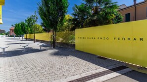 A paved walkway with a yellow fence labeled 'MUSEO CASA ENZO FERRAR' and a metal bench. - Museo Enzo Ferrari|Stazione FS-WiFi-parking NO ZTL (Modena)