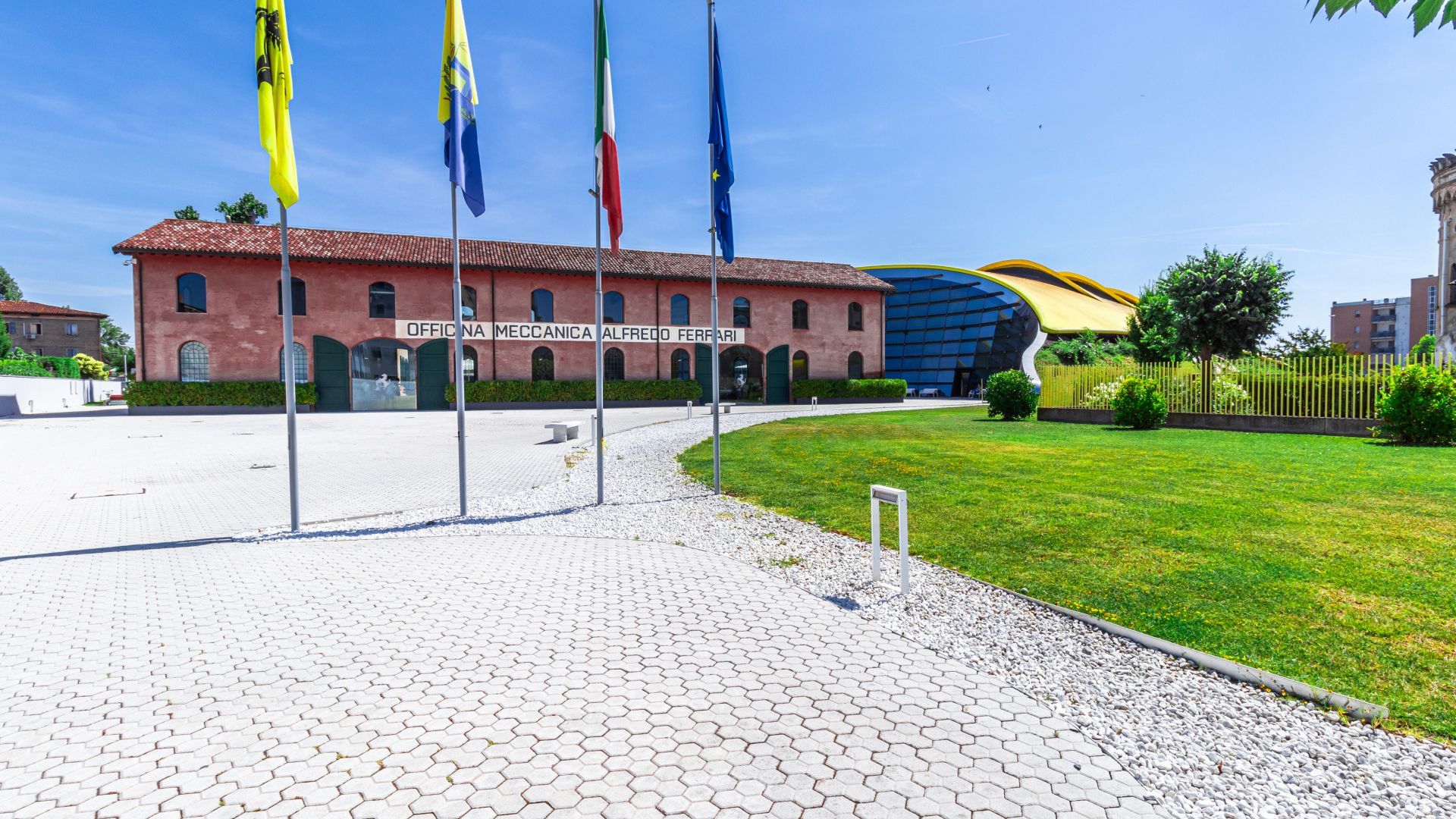 Ein gepflasterter Eingangsbereich, im Hintergrund ein Gebäude mit der Aufschrift 'OFFICINA MECCANICA ALFREDO FERRARI'.