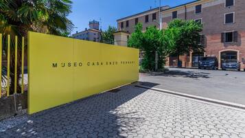 Un bâtiment jaune avec une enseigne indiquant « MUSEO CASA ENZO FERRARI », un chemin piétonnier pavé et une rue bordée de voitures garées, avec d’autres bâtiments en arrière-plan.