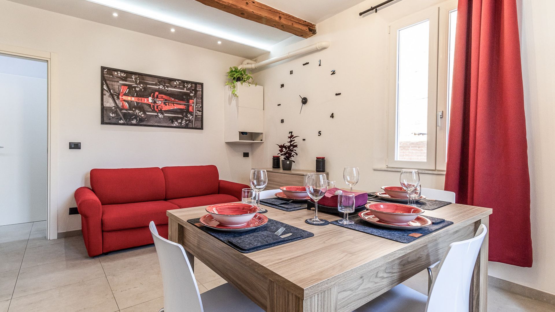 Un coin repas avec une table en bois dressée pour quatre personnes, des coussins rouges sur un canapé et une horloge murale.