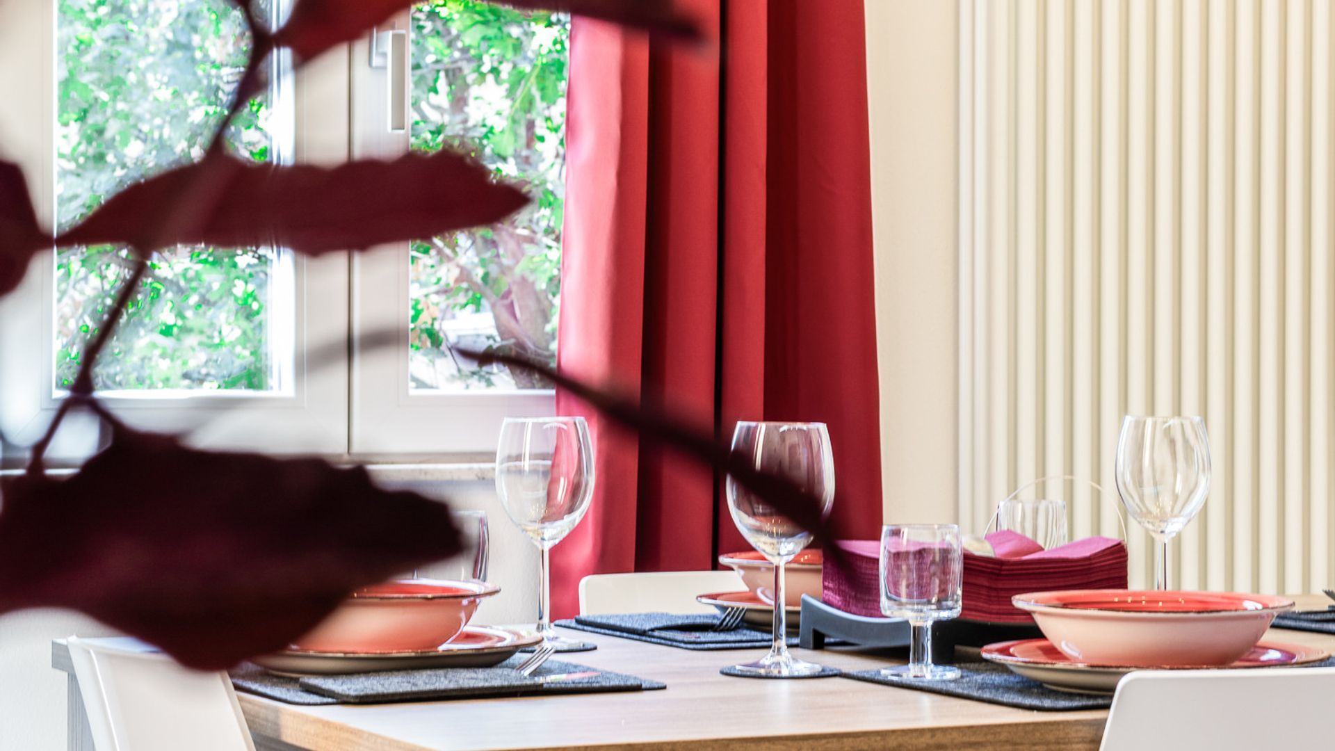 A dining table set with plates, glasses, and cutlery, with a window and curtains in the background.