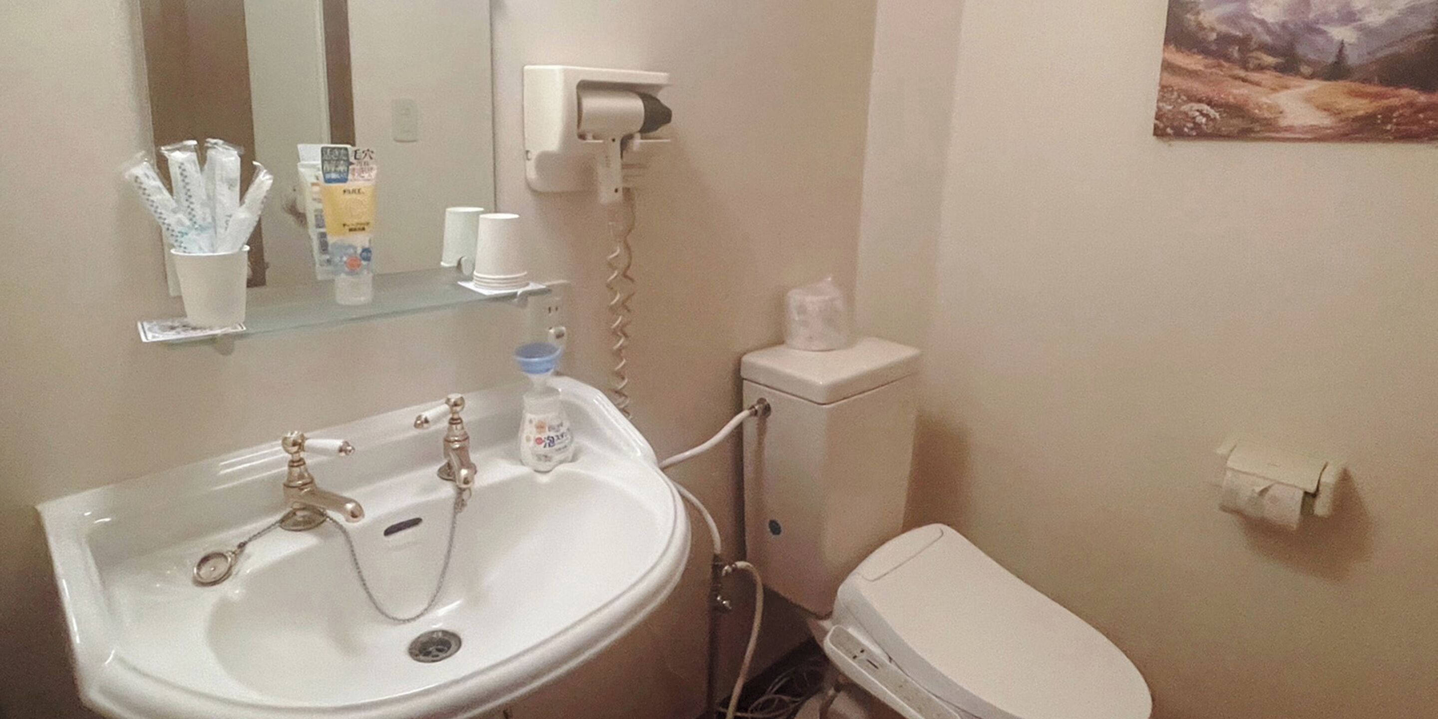 A bathroom with a white sink, mirror, and toilet. There are toiletries on the sink and a paper towel dispenser on the wall.