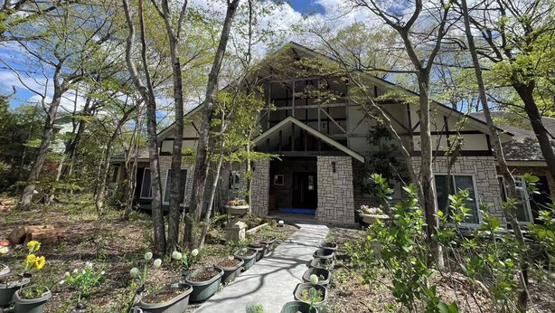 Una casa a due piani con facciata in pietra, un patio in legno e un giardino con vasi di piante e fiori.