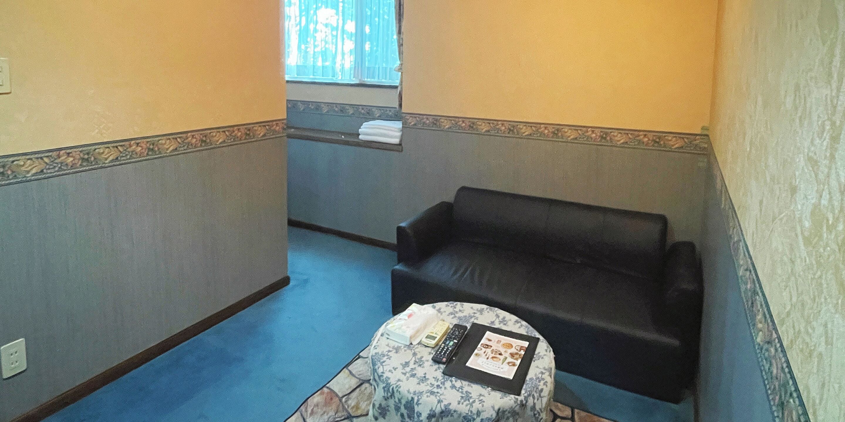 Ein kleiner, schwach beleuchteter Raum mit einer schwarzen Ledersofa, einem kleinen runden Tisch mit einer Fernbedienung und einer Speisekarte und einem Fenster mit Jalousien.
