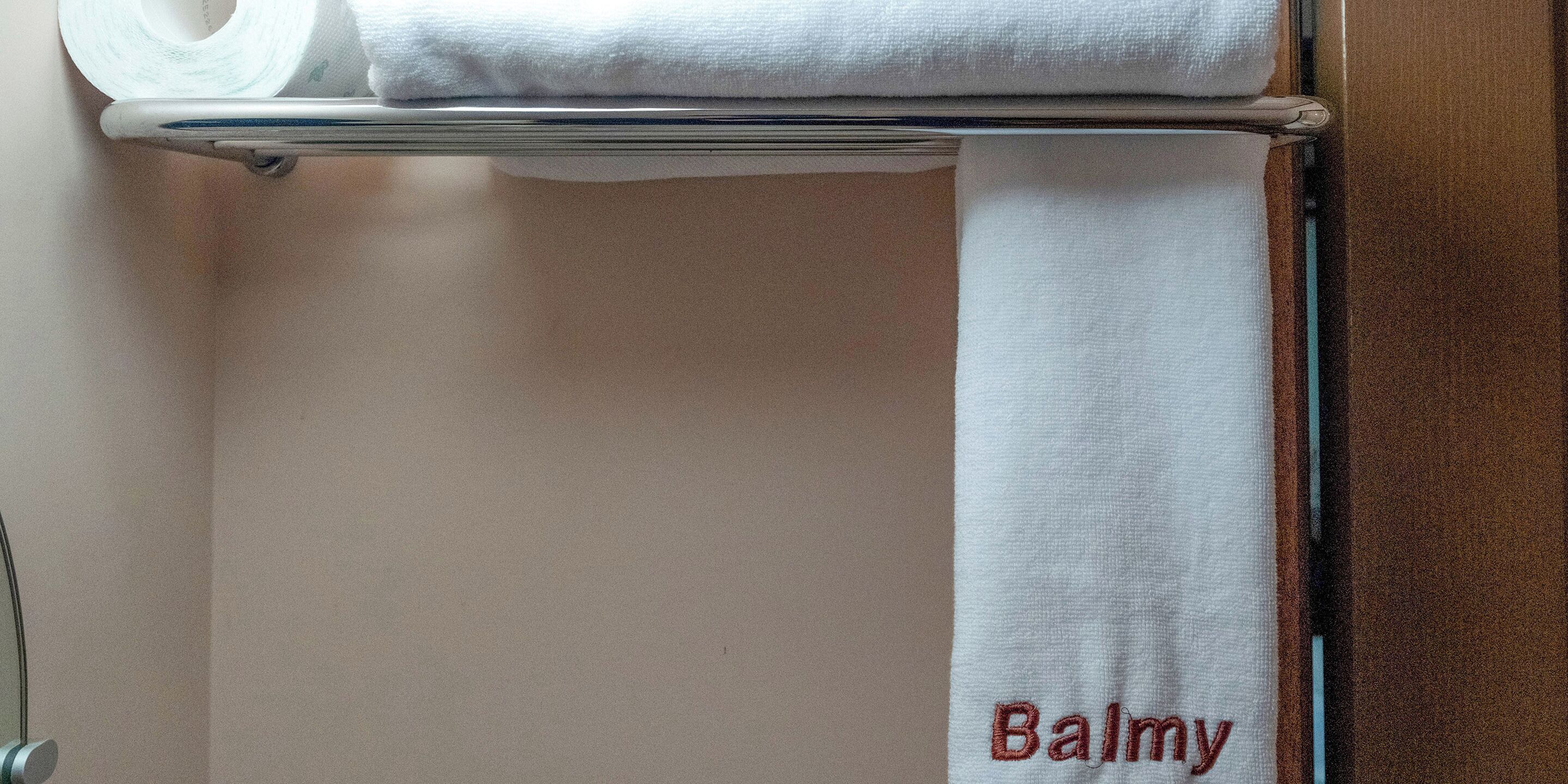 A bathroom with a towel rack holding a rolled towel and a towel with the name "Balmy" embroidered on it.