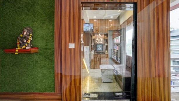 A hotel entrance with a wooden facade and a green artificial grass wall. Inside, there is a reception desk and a television.
