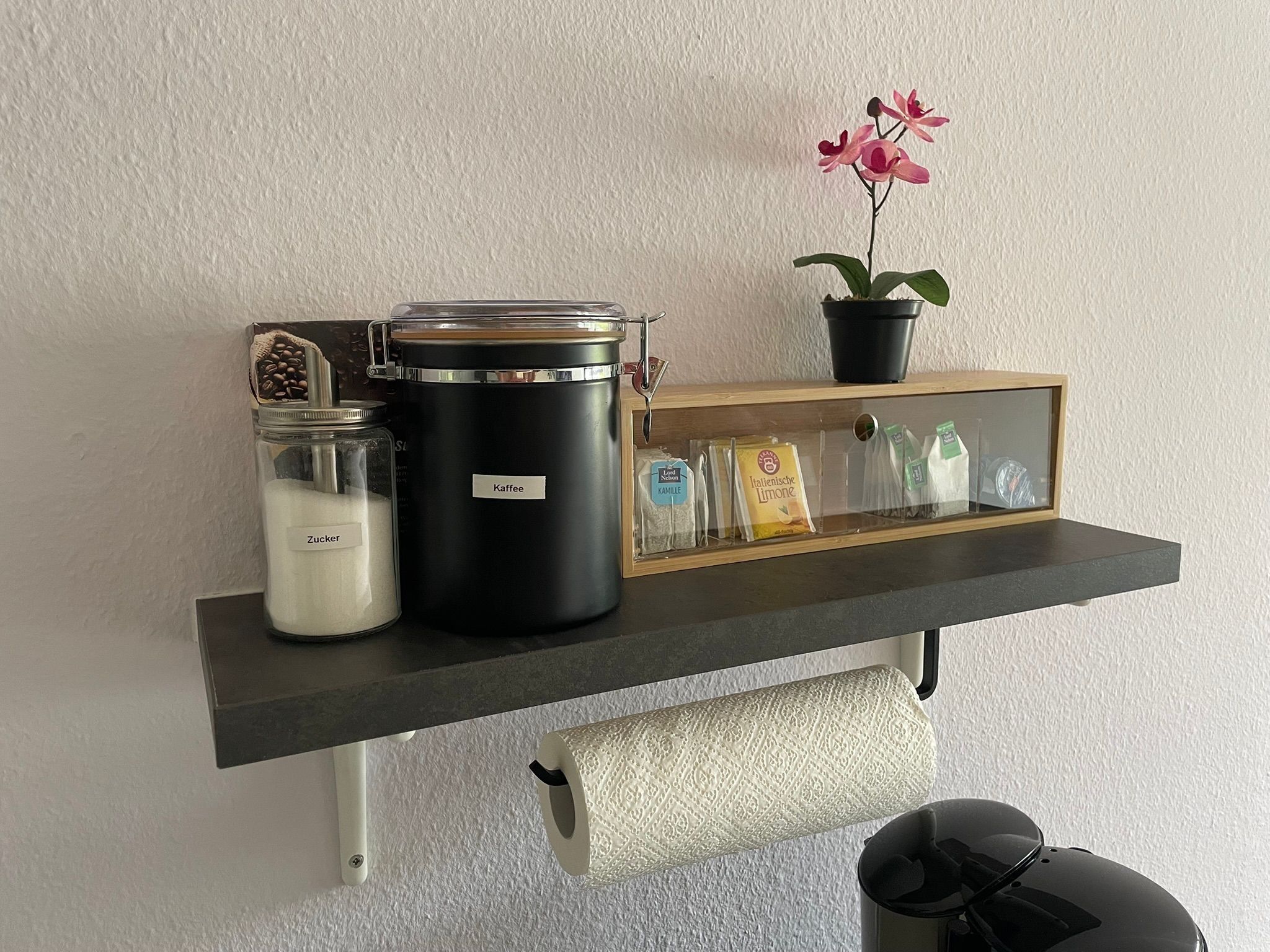 Eine Küchenarbeitsplatte mit Kaffeemaschine, Zuckerglas, einem schwarzen Behälter mit der Aufschrift 'Kaffee', einer Topfpflanze und einem Regal mit verpackten Lebensmitteln.