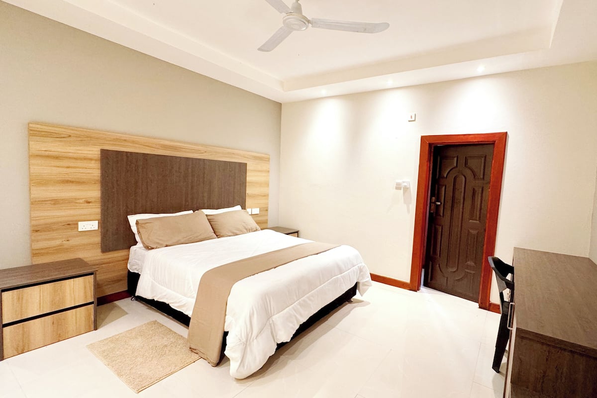 A bedroom with a bed, bedside tables, a ceiling fan, a door, and a small desk area.