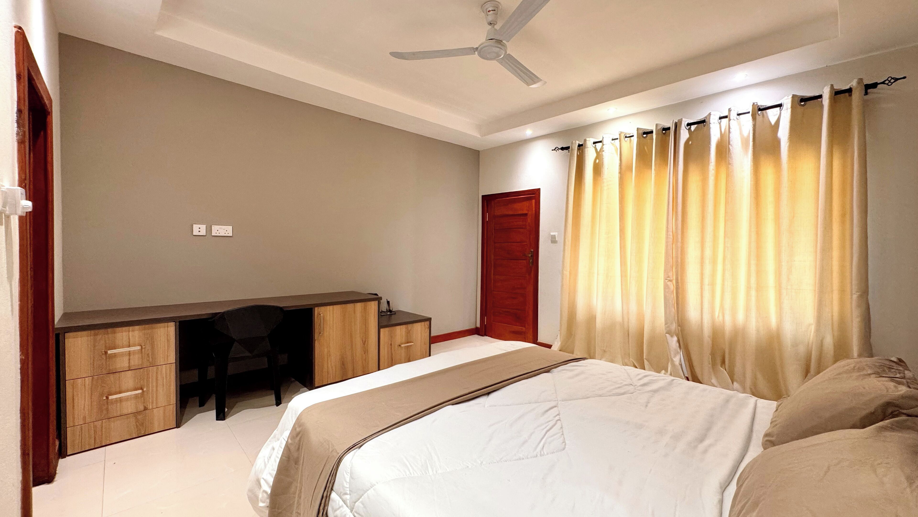 A bedroom with a bed, a desk, and a chair. There is a ceiling fan, a door, and curtains.