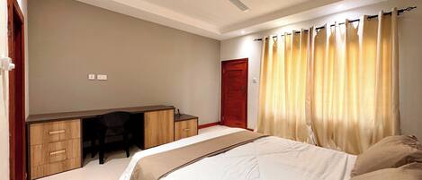A bedroom with a bed, a desk, and a chair. There is a ceiling fan, a door, and curtains.