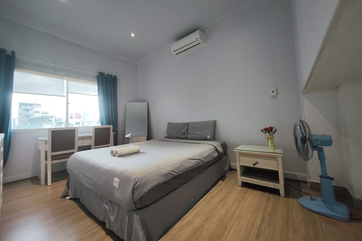 A modern hotel room with a large bed, a desk, a chair, a small table with a vase, and a standing fan.