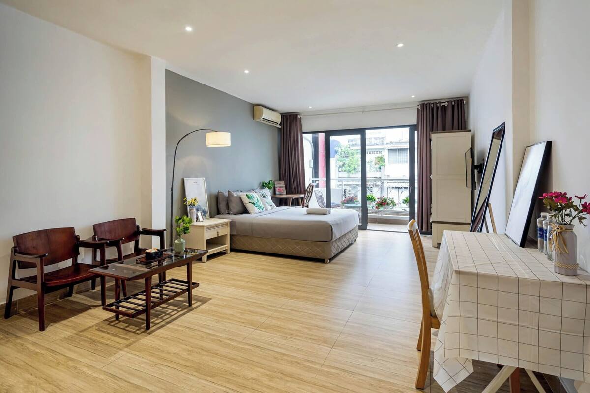A modern hotel room with a bed, a dining table, two chairs, a small table with a vase, and a large window offering a view of the city.