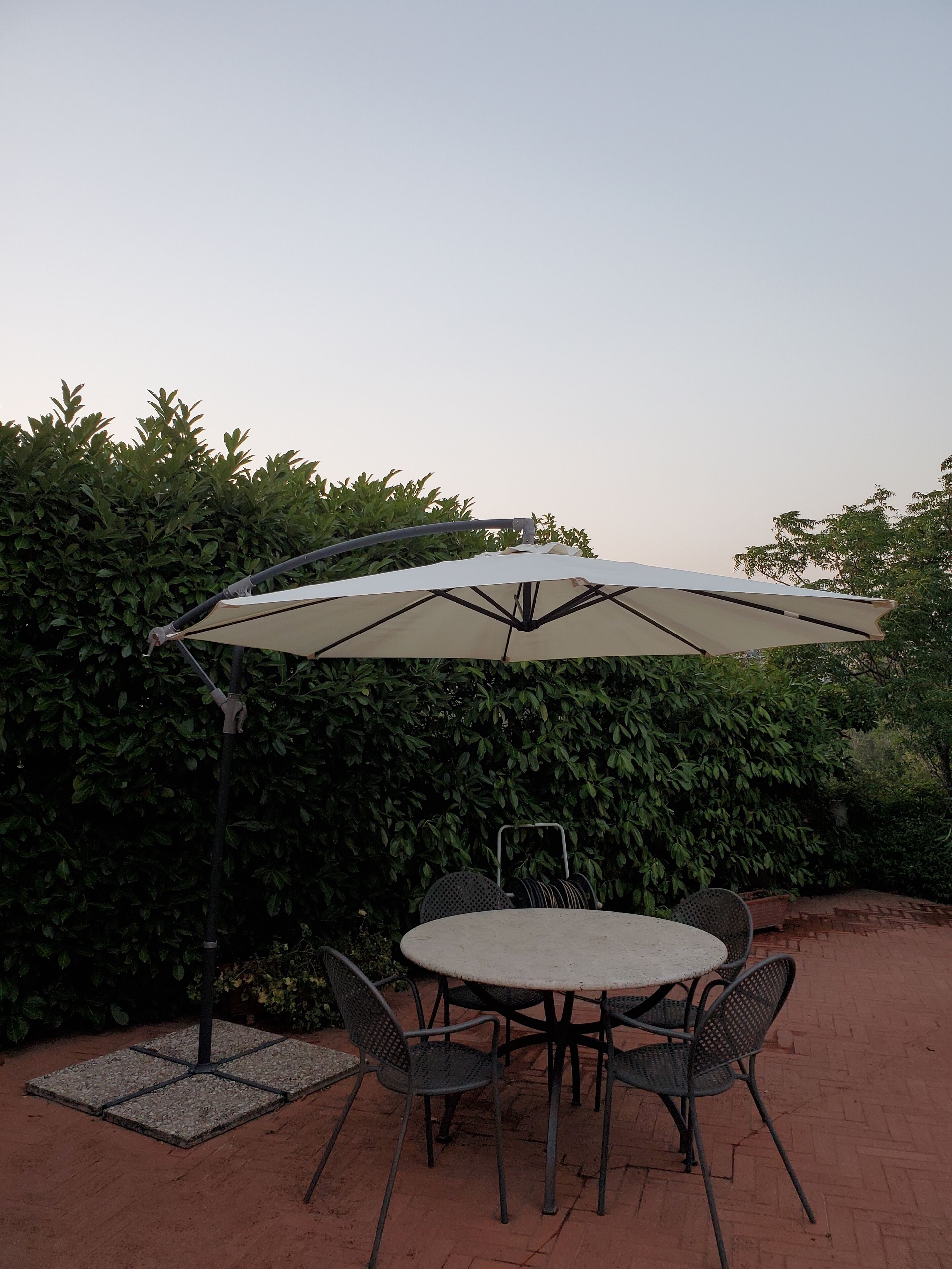 Een terras buiten met een ronde tafel, vier stoelen, een parasol en een metalen stoel met rugleuning.