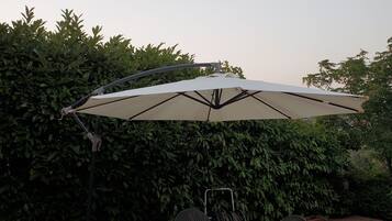 Een terras buiten met een ronde tafel, vier stoelen, een parasol en een metalen stoel met rugleuning.