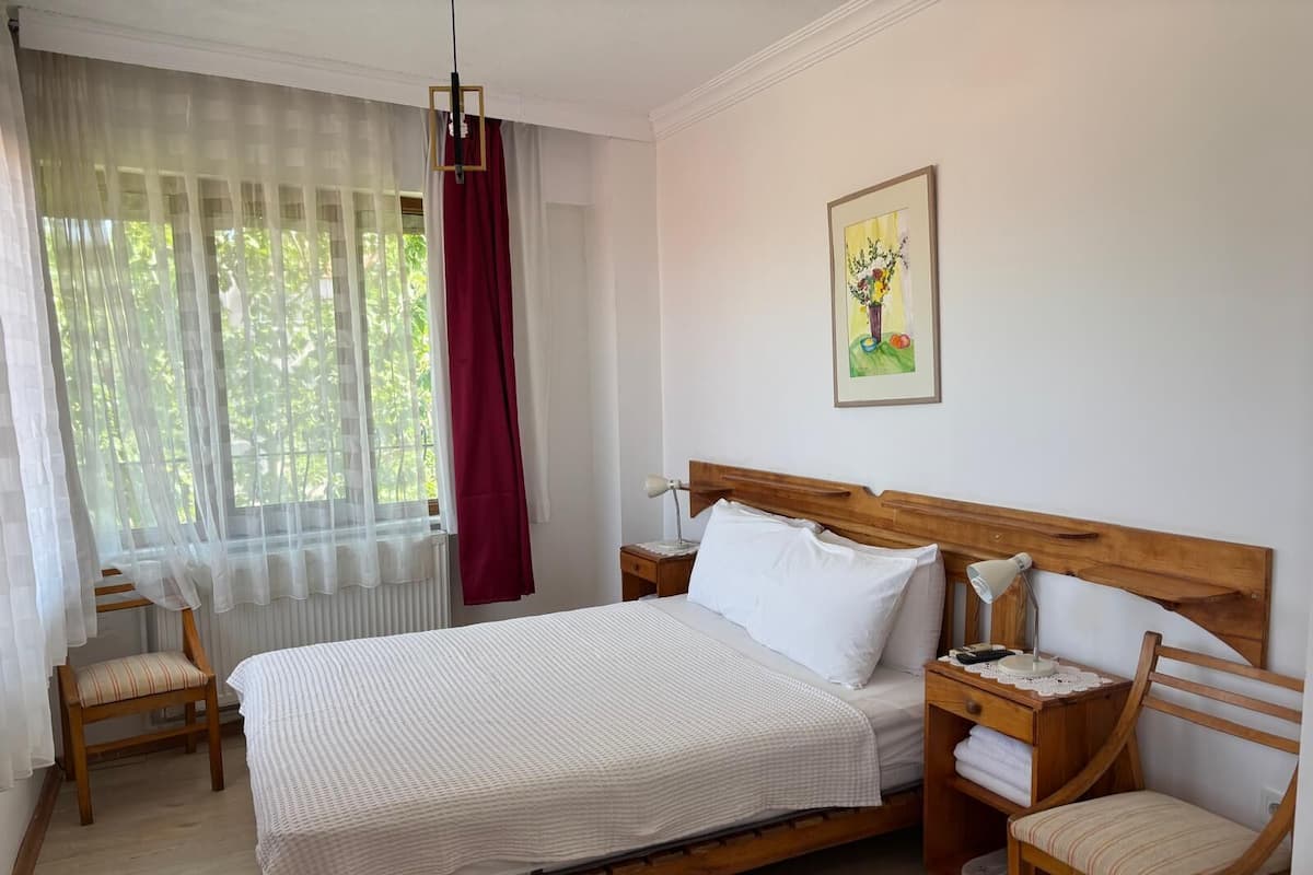 Un dormitorio ordenado con una cama de madera, ropa de cama blanca, una mesita de noche con lámpara, una silla y una ventana con cortinas translúcidas y un adorno rojo.