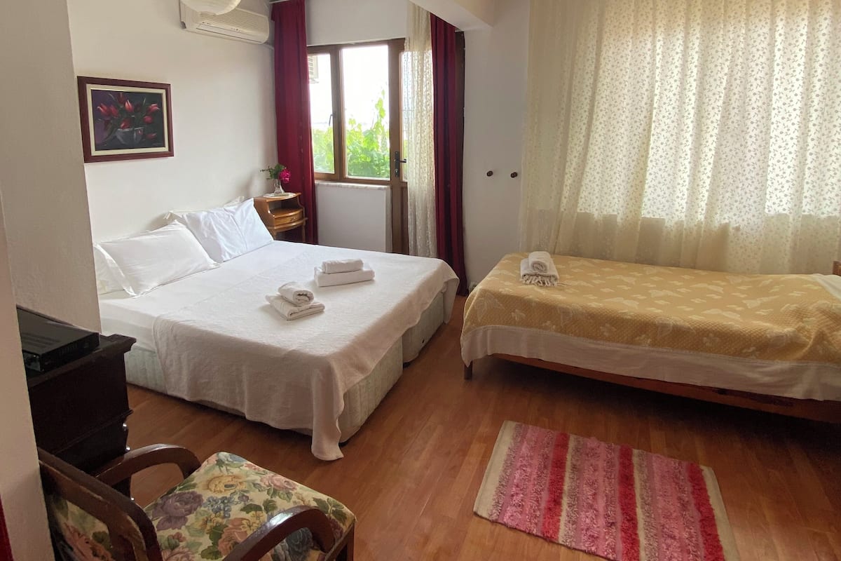 Habitación de hotel con dos camas, un escritorio, una silla, un ventanal con cortinas y una lámpara colgante.