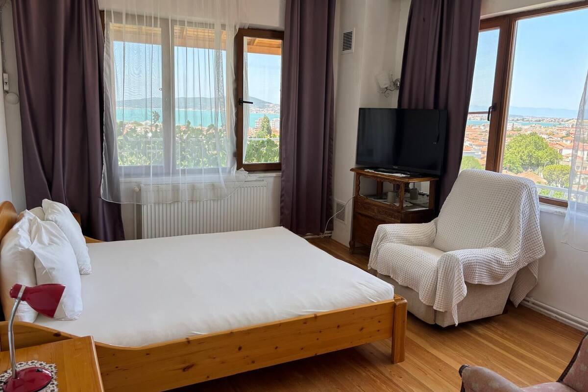 Un dormitorio con cama de madera, televisor sobre un mueble de madera, sillón blanco y vista al mar a través de amplias ventanas.