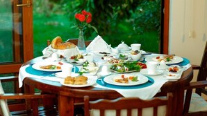 A round wooden table with a white tablecloth, surrounded by wooden chairs. The table is set with plates, bowls, cups, and a vase with red flowers. - Günebakan Taliani Otel (Ayvalik)