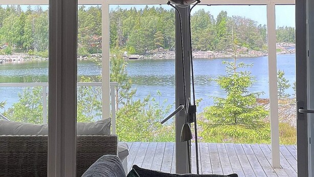 Una habitación con un amplio ventanal que da a una terraza con vistas a un lago, un sofá de mimbre y una lámpara de pie de metal.