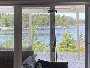 A room with a large glass door leading to a deck overlooking a lake, a wicker sofa, and a metal floor lamp. - 5 Star Holiday Home in Vaddo (Väddö)