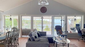 Una habitación amplia con techo alto, un candelabro, un gran ventanal con vista a un lago, un área de comedor con sillas y una mesa, y un rincón de estar con un sofá y sillones.