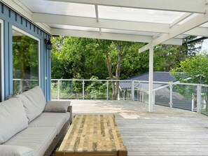 A covered outdoor deck with a sofa and a wooden table, surrounded by a white railing and trees in the background. - 5 Star Holiday Home in Vaddo (Väddö)