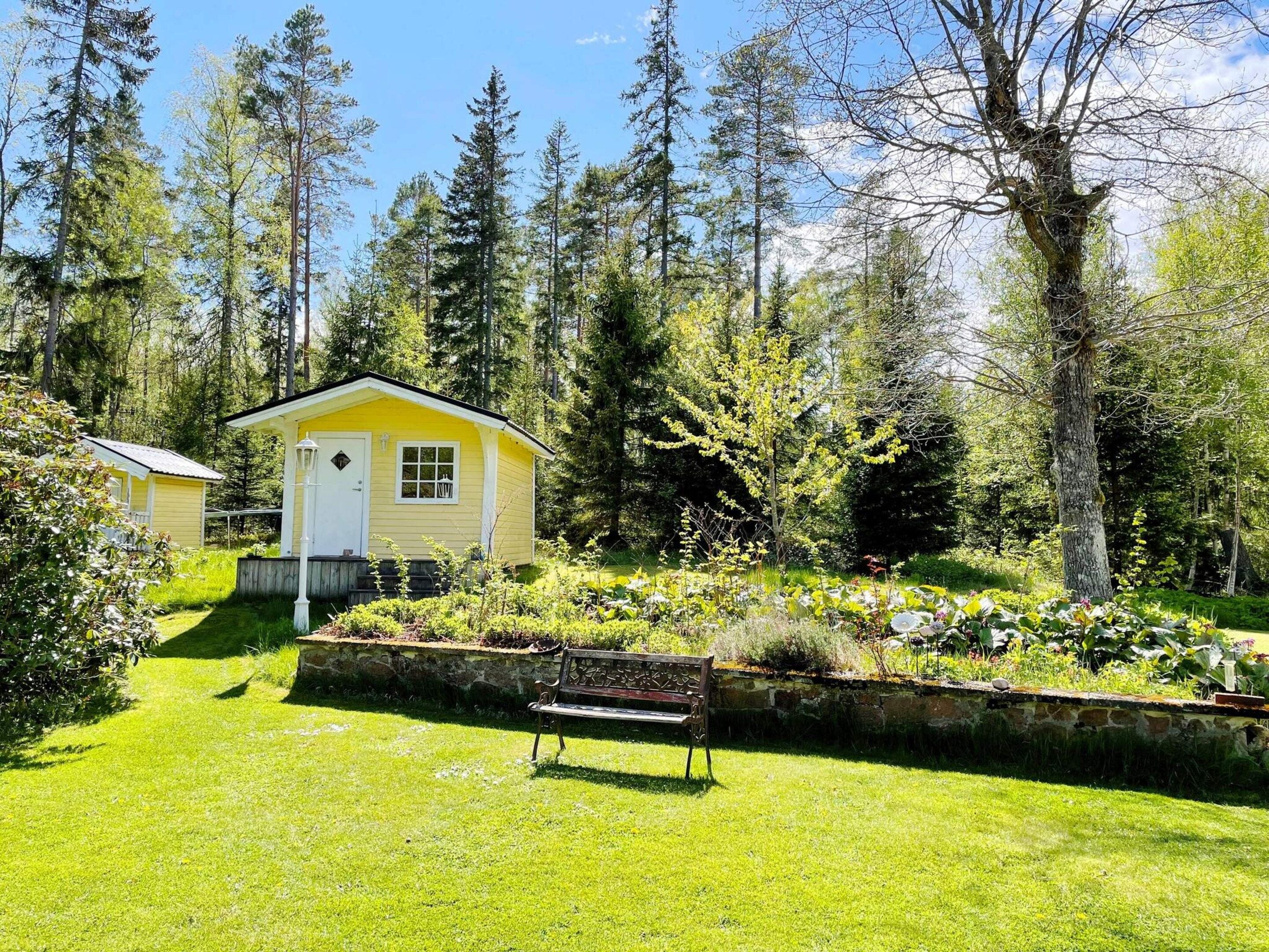 Una cabaña pequeña y amarilla con puerta blanca, rodeada de un jardín cuidado y altos árboles.