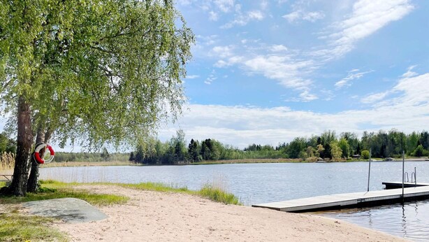 Eine Seenlandschaft mit Sandstrand, einer anlegenden Mole und einem Rettungsring, der an einem Baum hängt.
