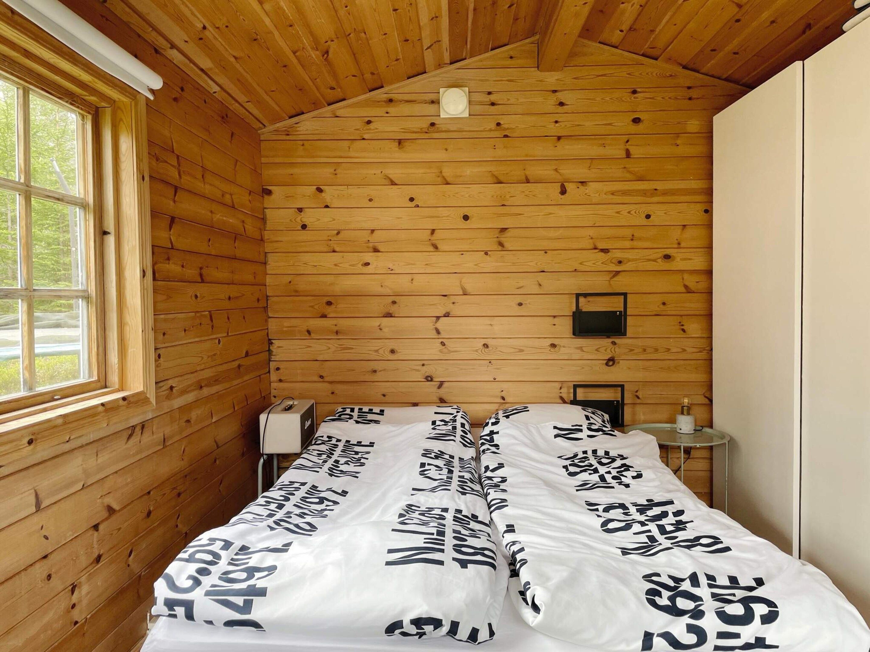 A small, single-bed room with wooden walls and a sloped ceiling. There is a window on the left side, a bedside table with a lamp, and a small shelf with a few items.