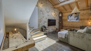 A rustic interior with a stone wall, wooden beams, a wooden staircase, a flat-screen TV, a woven light fixture, and a sofa. - 🏡 La Cabane à Camillo – Warm and Cozy Retreat (CHEVALINE)