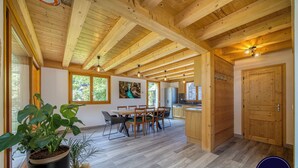 A spacious room with a wooden ceiling, a dining area with a table and chairs, a kitchen with a refrigerator, and a large window with a view of greenery. - 🏡 La Cabane à Camillo – Warm and Cozy Retreat (CHEVALINE)