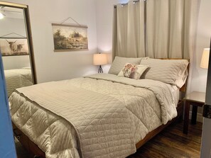 A bedroom with a bed, a bedside table, a lamp, a mirror, and a wall-mounted picture. - 🌵 “Mountain View 2BR Country Home -Netflix – Spacious (Hesperia)