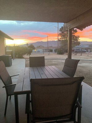 An outdoor patio with a wooden table and chairs, a covered area, and a view of a sunset over a distant landscape. - 🌵 “Mountain View 2BR Country Home -Netflix – Spacious (Hesperia)