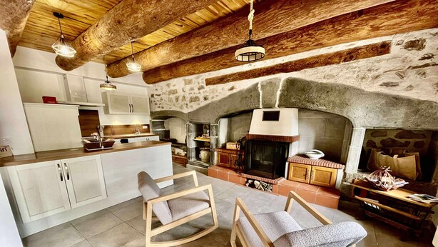 Une cuisine rustique avec un mur en pierre, des poutres en bois et une cheminée. On y trouve deux chaises en bois et une petite table.