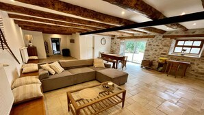 Un vaste espace de vie comprenant un grand canapé d’angle, une table basse en bois et une table à manger avec des chaises. La pièce est agrémentée de poutres apparentes en bois au plafond et de murs en pierre.