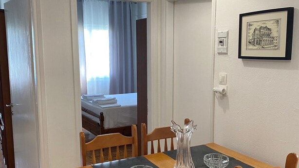 A dining area with a wooden table and chairs, a hanging light fixture, a framed picture on the wall, and a bed visible through an open door. - Corfu Town Flat (Kerkira)