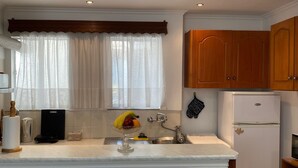 A compact kitchen with a central island, wooden cabinets, a refrigerator, and a window with sheer curtains. - Corfu Town Flat (Kerkira)