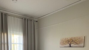 A bedroom with a bed, a nightstand with a lamp, a window with curtains, and a wall-mounted painting. - Corfu Town Flat (Kerkira)