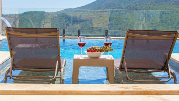 Zwei Liegestühle, zugewandt zu einem Pool, dazwischen ein Tisch mit Obstschale und zwei Gläsern Rotwein.