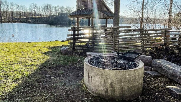 En træterrasse med et bålsted og en lille træbygning ved en sø.