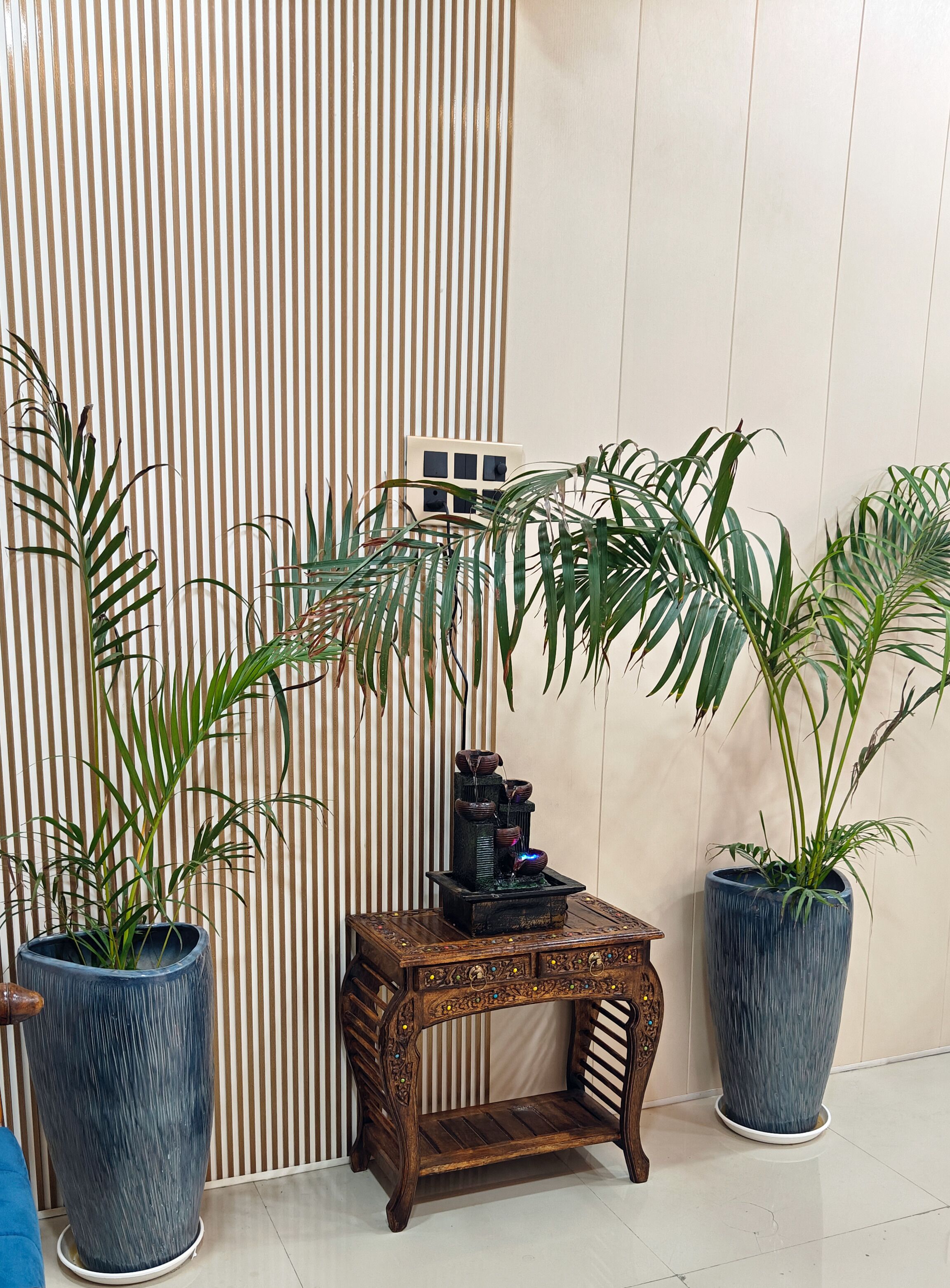 A corner with a striped wall, a small wooden table with a decorative fountain, and two large potted plants.