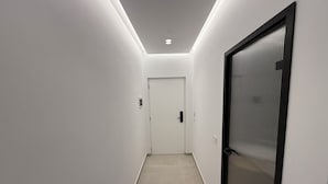 Un pasillo estrecho y bien iluminado, con una puerta al final, una pared texturizada a la derecha y un interruptor de luz a la izquierda.