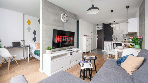 Ett modernt vardagsrum med en grå soffa, en tv-skärm som visar "NETFLIX", ett skrivbord med en bärbar dator och en matplats i bakgrunden.